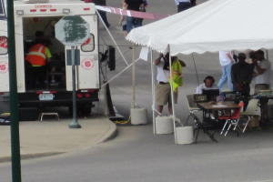 Ein Krankenwagen steht auf der linken Seite einer Straße, während auf der rechten Seite ein Zelt eine Gruppe von Menschen schützt, mit einem Tisch und einem Stuhl in der Nähe.