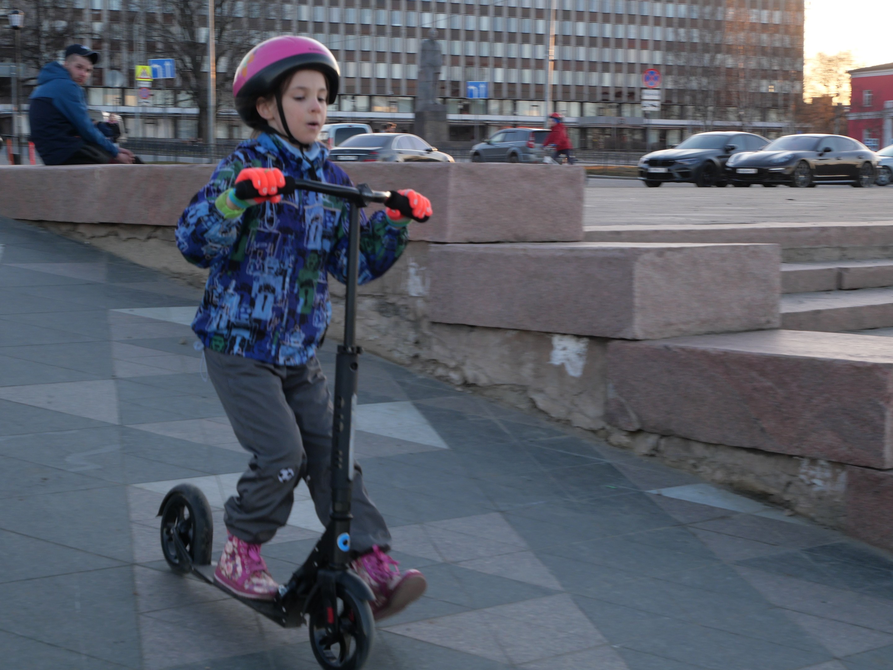Ein junger Junge in einem Helm und Handschuhen fährt auf einem Roller eine Treppe auf dem Gehweg hinunter, mit Fahrzeugen, Menschen, Bäumen, Polen, Brettern, Gebäuden und einem klaren blauen Himmel im Hintergrund.