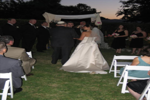 Eine Gruppe von Menschen bei einer Hochzeit, wobei die Frauen rechts Blumensträuße halten, während andere auf Stühlen sitzen und Bäume im Hintergrund zu sehen sind.