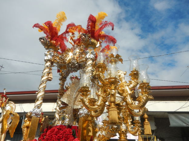 Ein großer goldener und roter Float, der mit Blumen und anderen Dekorationsgegenständen geschmückt ist, in einem Karnevalsumzug, mit einem Gebäude, Strommasten mit Drähten und einem bewölkten Himmel im Hintergrund.