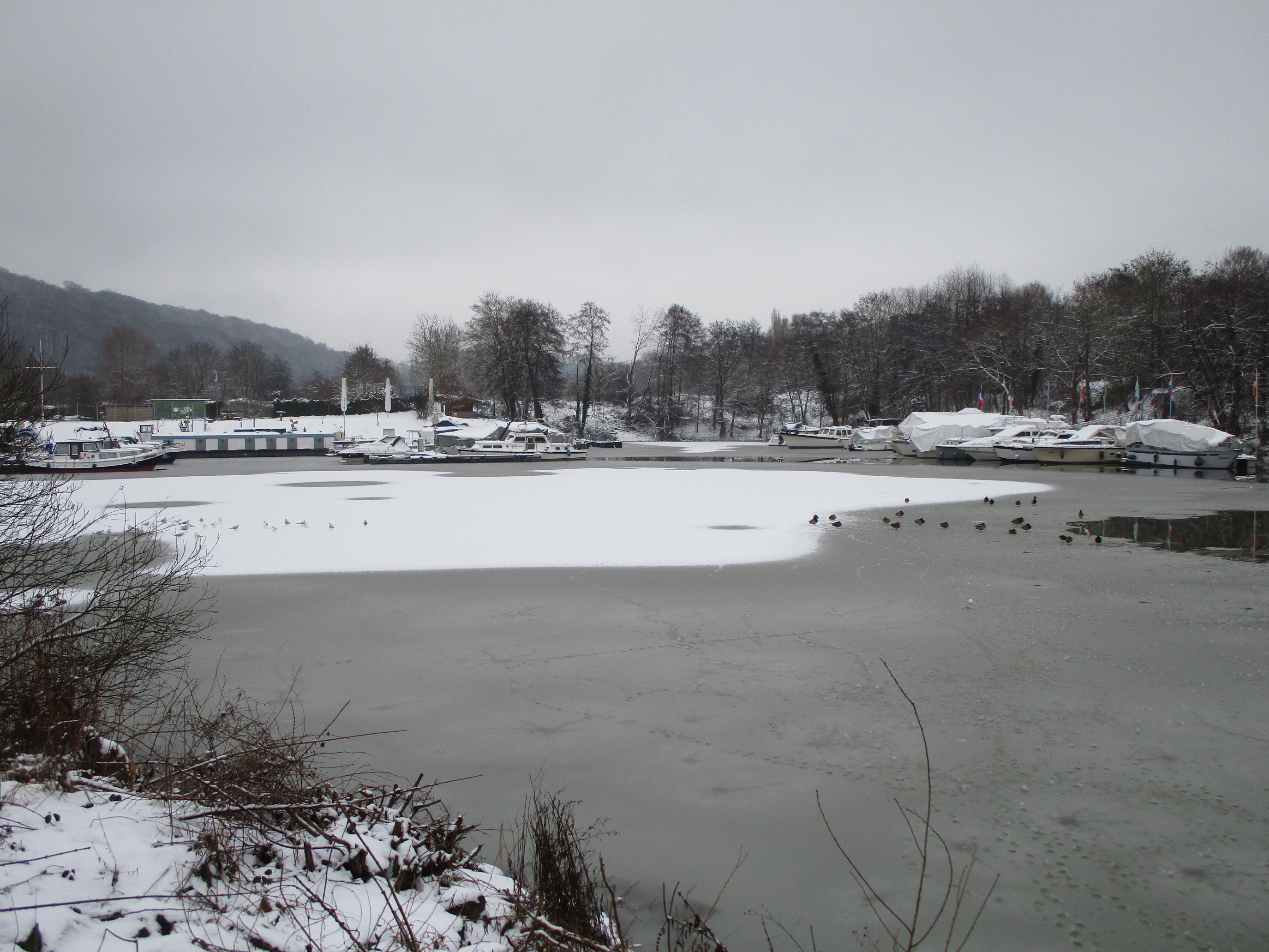 Ein zugefrorener See mit Booten im Wasser, umgeben von schneebedeckten Pflanzen und Bäumen, mit Hügeln und einem klaren Himmel im Hintergrund.