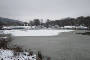Ein zugefrorener See mit Booten im Wasser, umgeben von schneebedeckten Pflanzen und Bäumen, mit Hügeln und einem klaren Himmel im Hintergrund.