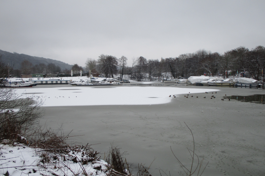 Ein zugefrorener See mit Booten im Wasser, umgeben von schneebedeckten Pflanzen und Bäumen, mit Hügeln und einem klaren Himmel im Hintergrund.