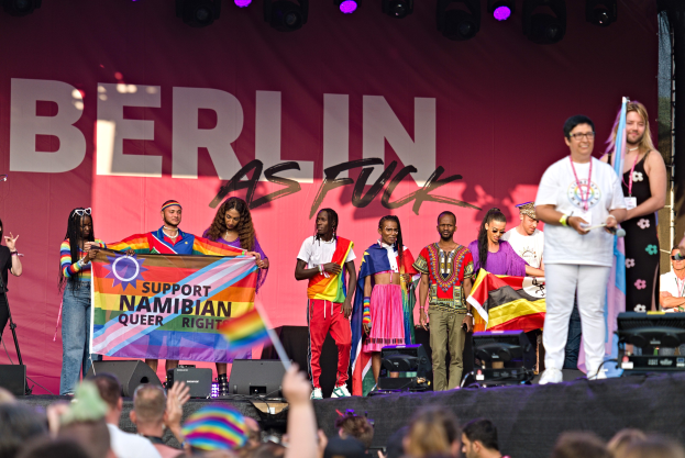 Menschen auf einer Bühne mit einem Banner 'Berlin as Fuck' und Lautsprechern, Mikrofonen und einer jubelnden Menge unten.