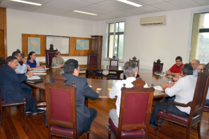 Eine Gruppe von Menschen, die an einem Tisch mit Papieren und Gläsern sitzt, ein Klimagerät, ein Anschlagbrett und Karten an der Wand im Hintergrund.