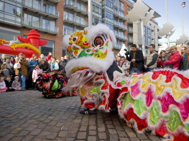 Ein lebendiger chinesisches Neujahrsfest in Amsterdam mit einer Löwen-Tanzvorstellung vor einer Zuschauermenge, darunter einige, die das Ereignis fotografieren, vor einer Kulisse aus Gebäuden, Laternenmasten und einem klaren blauen Himmel.