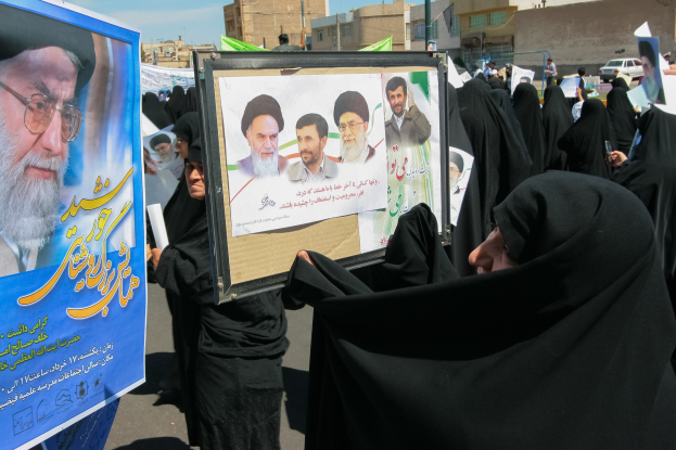 Eine Gruppe von Frauen in schwarzen Abayas hält Plakate mit Fotos von Ayatollah Ali Khamenei vor einem Hintergrund aus Gebäuden, Fahrzeugen und einem klaren blauen Himmel.