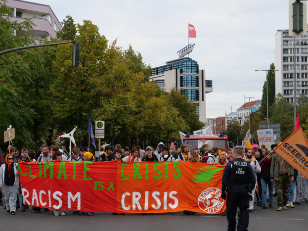 Eine Gruppe von Menschen marschiert mit einem Banner, auf dem "Klima-Krise ist eine Krise" steht, eine Straße entlang, umgeben von Bäumen, Laternenmasten und Schildern, einem geparkten Fahrzeug, Gebäuden und einem klaren blauen Himmel.