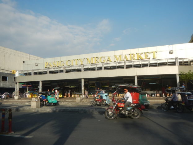 Ein belebter Pasig City Mega Market mit Fahrzeugen auf der Straße, Menschen, die herumlaufen, einem Gebäude mit Text im Hintergrund und einer bewölkten Himmel.