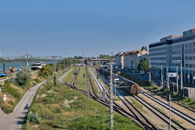 Ein Zug fährt auf Schienen neben einer Stadtlandschaft mit Strommasten, Gebäuden, Bäumen, Fahrzeugen, einer Brücke, Wasser und einer bewölkten Himmel.