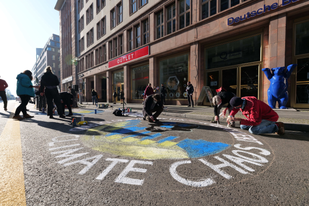 Menschen sitzen vor einem Gebäude mit Fenstern und Namensschildern auf dem Boden, umgeben von Flaschen und anderen Gegenständen, während sie an einer Klimawandel-Demonstration in Berlin mit Bäumen und einem klaren blauen Himmel teilnehmen.