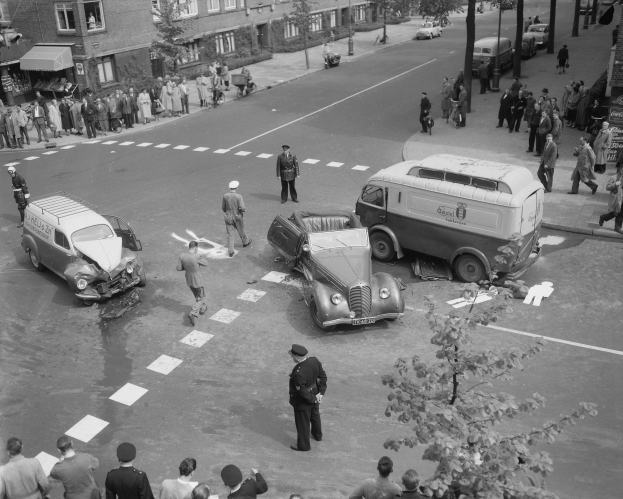 Schwarz-weiß-Szene eines Autounfalls an einer Straßenkreuzung mit mehreren Fahrzeugen, darunter ein Lieferwagen, und einer Gruppe von Schaulustigen; Gebäude, Bäume, Strommasten und Schilder sind im Hintergrund zu sehen.
