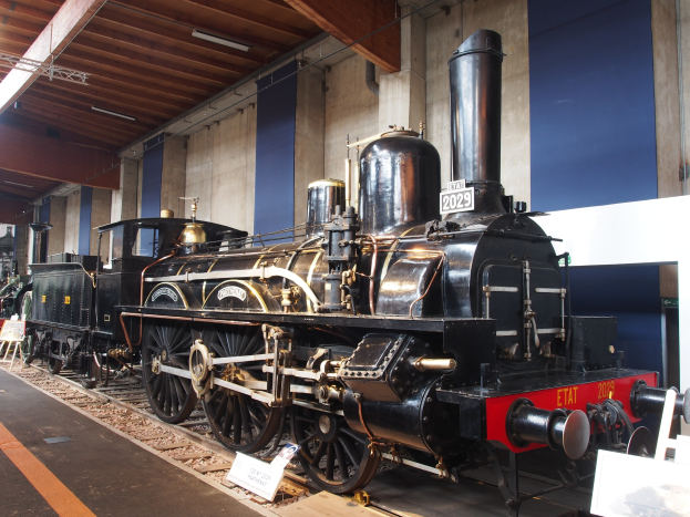 Eine große, alte Dampflokomotive in einem Museums Setting umgeben von Eisenbahnschienen, Informationsschildern, Ständen und anderen Ausstellungsstücken, mit einer Wand- und Deckenbeleuchtung im Hintergrund.