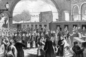 Schwarz-weißes Bild einer überfüllten Bahnhofshalle mit Menschen in verschiedener Kleidung, einem Zug auf den Schienen im Hintergrund, hängenden Lampen links und einer Person mit einer Kiste rechts.