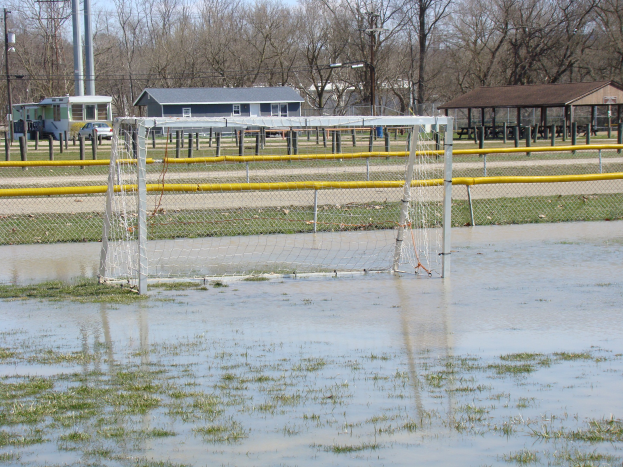 Ein Fußballtor steht in einem überfluteten Feld mit umliegender Grasfläche und Wasser, mit Hütten, Pfählen, Bäumen, Fahrzeugen und einem klaren blauen Himmel im Hintergrund.