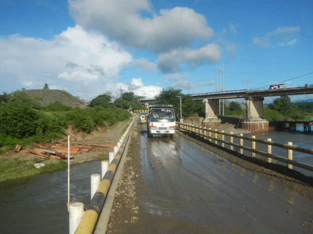 Ein Lastwagen fährt durch Überschwemmungswasser neben einer Brücke mit Geländern, mit Bäumen, Strommasten und einem bewölkten Himmel im Hintergrund.