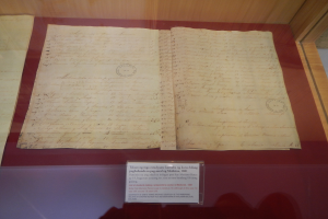 Altes Dokument in einer Glasvitrine mit Text darauf und einer Beschriftung unten, vor einem hölzernen Hintergrund in einem Museum.