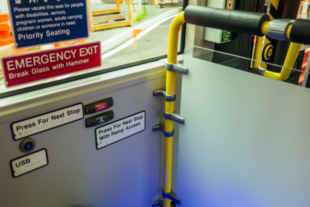 Notausgangsschild an der Seite eines Zuges mit einer Tür, Griff und einem Glasfenster, das Straße, Pfosten und andere Objekte draußen zeigt.