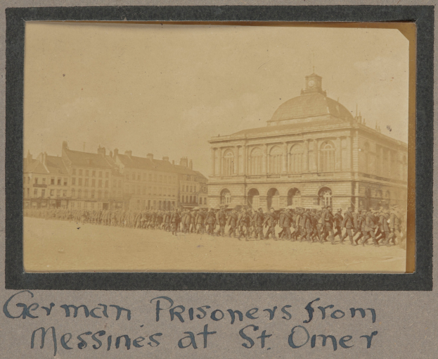 Eine Gruppe deutscher Kriegsgefangener aus Messines steht vor einem Geb"ude unter einem klaren blauen Himmel, mit Text am unteren Bildrand.