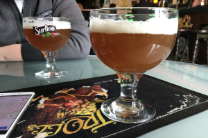 Zwei Gläser goldenes Bier auf einem Tisch mit einem Handy in der Nähe, in einem Raum mit gerahmten Wanddekoration und Menschen im Hintergrund.