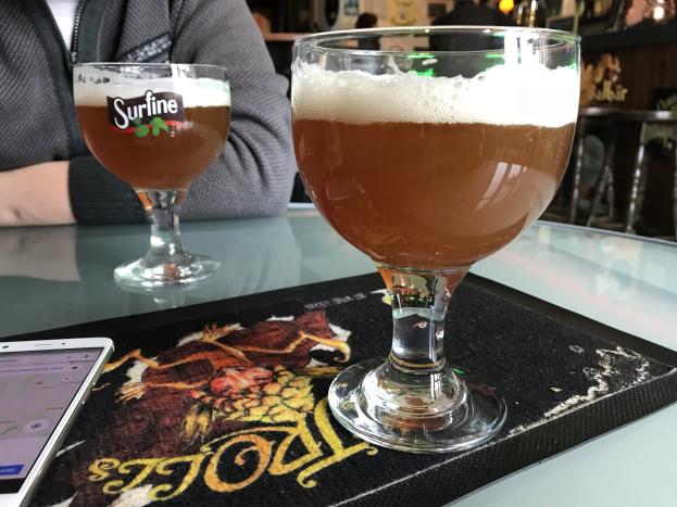 Zwei Gläser goldenes Bier auf einem Tisch mit einem Handy in der Nähe, in einem Raum mit gerahmten Wanddekoration und Menschen im Hintergrund.