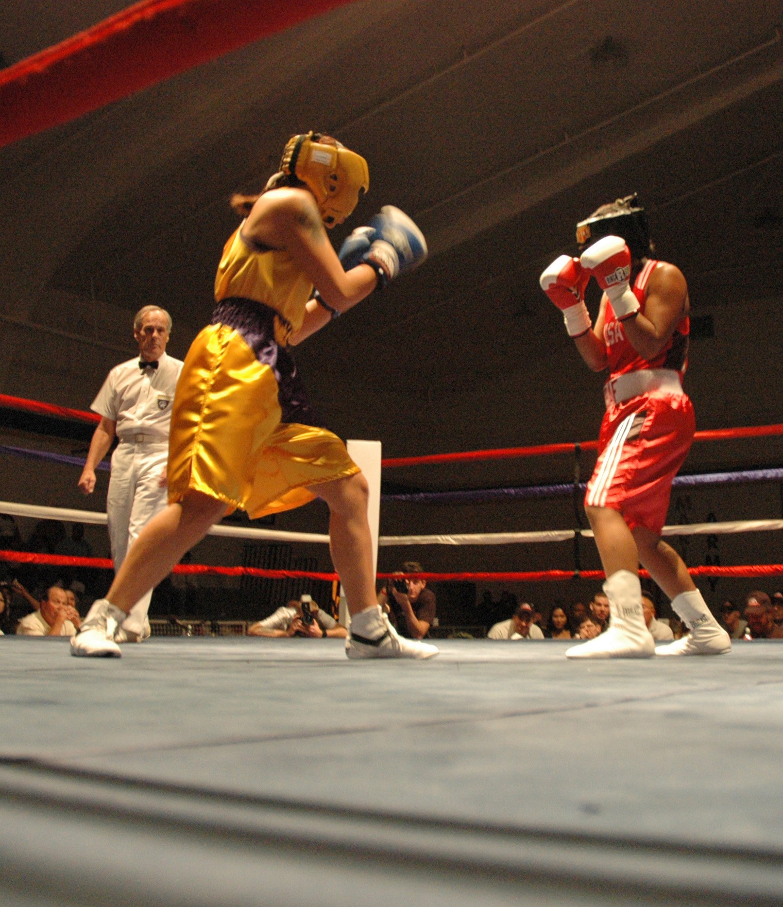Zwei Boxer im Ring mit einem Schiedsrichter und Zuschauern, einige halten Kameras, unter einer sichtbaren Decke.