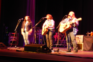 Drei Frauen performen auf der Bühne eines Musikkonzerts, jede singt in ein Mikrofon und spielt eine Gitarre, mit einem Tisch voller Wasserflaschen und zusätzlichen Gitarren dahinter.