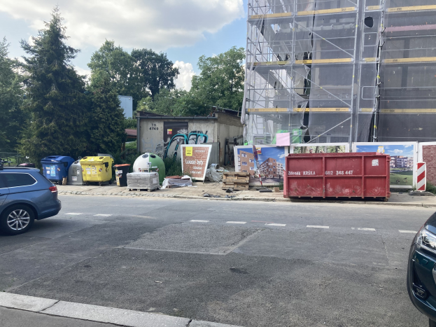 Zwei Autos neben einem Gebäude im Bau parken mit verstreuten Straßenobjekten, Bäumen und einem bewölkten Himmel im Hintergrund.