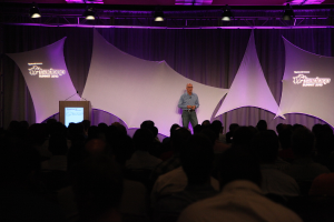 Ein Mann in einem blauen Hemd steht auf einer B├╝hne mit schwarzen und weißen Vorh├Ąngen, spricht zu einem sitzenden Publikum unter einem Metallrahmen mit einer Spotlight.