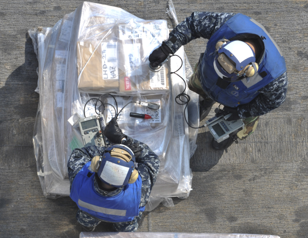 Zwei Männer in blauen Uniformen und Helmen arbeiten an einem großen Drohnenmodell, halten verschiedene Gegenstände und eine Plastikabdeckung liegt auf dem Boden.