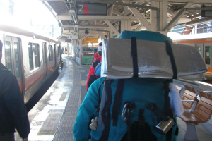 Ein Bahnhofsplatz mit Menschen neben einem Zug, einem Stand in der Mitte, einem weiteren Zug auf der rechten Seite und einer Bank in der rechten unteren Ecke.