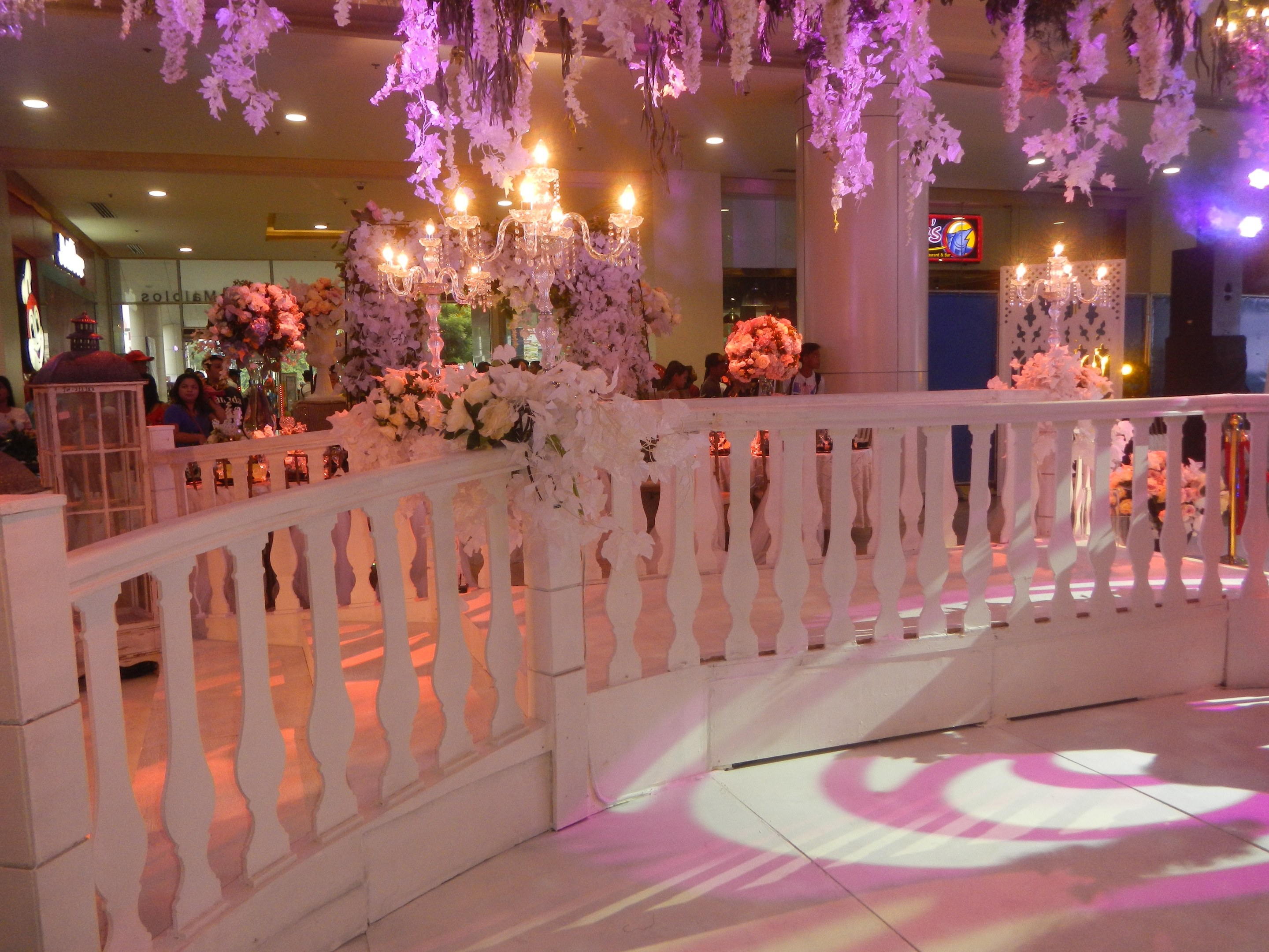 Weißes Geländer mit Blumen geschmückt und von der Decke hängende Kronleuchter, umgeben von Menschen, Säulen und Lichter im Hintergrund und ein Bildschirm auf der rechten Seite des Bildes.