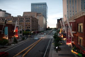 Stadtpanorama-Foto, das eine Stra├če mit Autos zeigt, flankiert von Geb├Ąuden und Wolkenkratzern, unter einem Himmel.