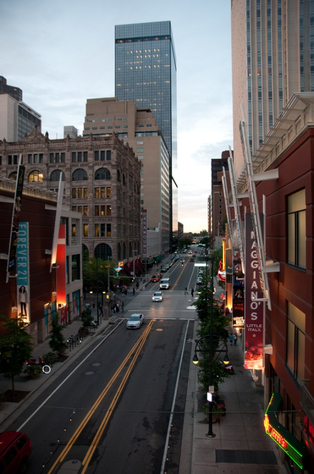 Stadtpanorama-Foto, das eine Stra├če mit Autos zeigt, flankiert von Geb├Ąuden und Wolkenkratzern, unter einem Himmel.