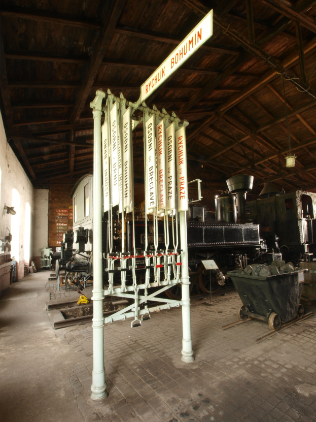 Ein Bahnsteig mit einem Zug auf den Gleisen, einem Rollstuhl auf dem Boden, einem Wegweiser, einer Wand auf der linken Seite, einer Decke oben und einer zentralen Signalbox.