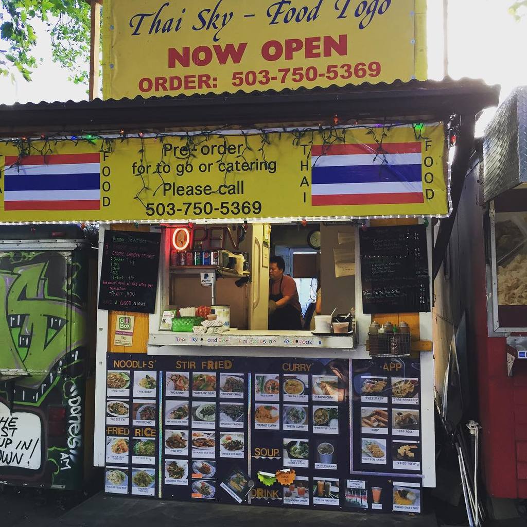 Ein Ladengeschäft mit verschiedenen Artikeln wie Getränkedosen, Speisekarten, einer Uhr, Gewürzen in Plastikbehältern, Gewürzen, Werbetafeln und Namensschildern unter dem Himmel.