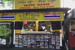 Ein Ladengeschäft mit verschiedenen Artikeln wie Getränkedosen, Speisekarten, einer Uhr, Gewürzen in Plastikbehältern, Gewürzen, Werbetafeln und Namensschildern unter dem Himmel.