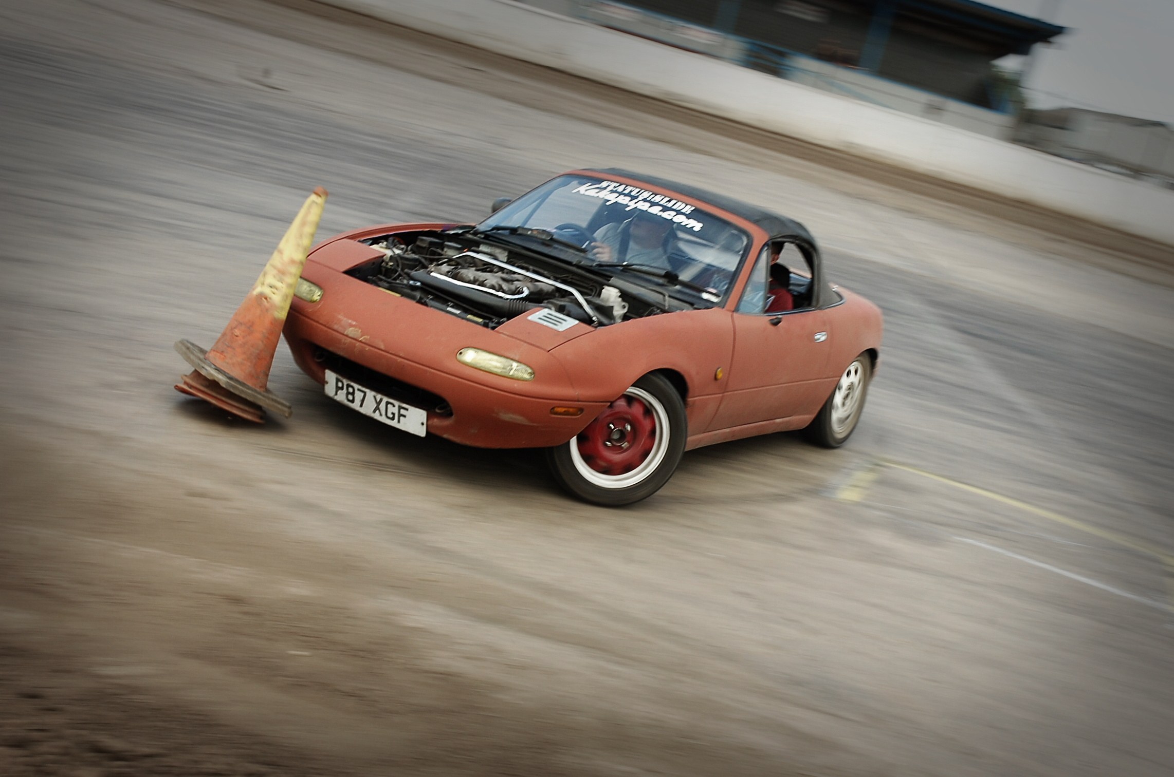 Eine Person fährt ein rotes Auto auf einer Rennstrecke mit einem Verkehrskegel vor sich, der Hintergrund ist durch hohe Geschwindigkeit unscharf.