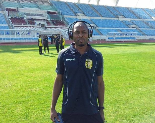 Ein Mann mit Headset, wahrscheinlich der Trainer der ghanaischen Nationalmannschaft, steht auf einem Fussballfeld in der Nähe eines Stadions und hält ein Objekt, während im Hintergrund Menschen zu sehen sind und der Himmel sichtbar ist.