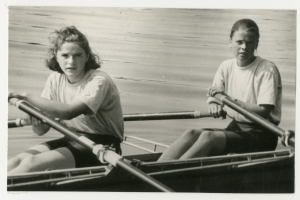 Zwei Frauen rudern ein schwarzes und weißes Boot auf ruhigem Wasser, halten Ruder in den Händen und scheinen zu rasen.