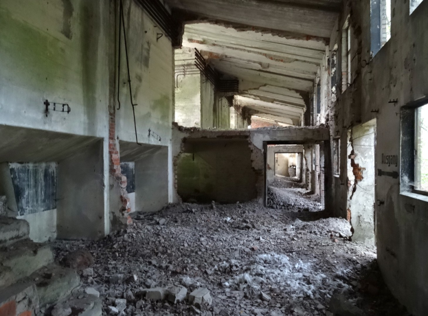 Altes Geb├Ąude mit umfangreichen Sch├Ąden, darunter eingestürzte W├Ąnde, zerbrochene Fenster, verstreute Trümmmer, freiliegende Rohre und Schutt über den Boden.