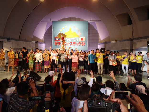 Eine Gruppe von Menschen auf einer Bühne, einige halten Kameras und Handys, andere Musikinstrumente, mit einem Banner und Lichtern im Hintergrund bei einer Festivaleröffnung.