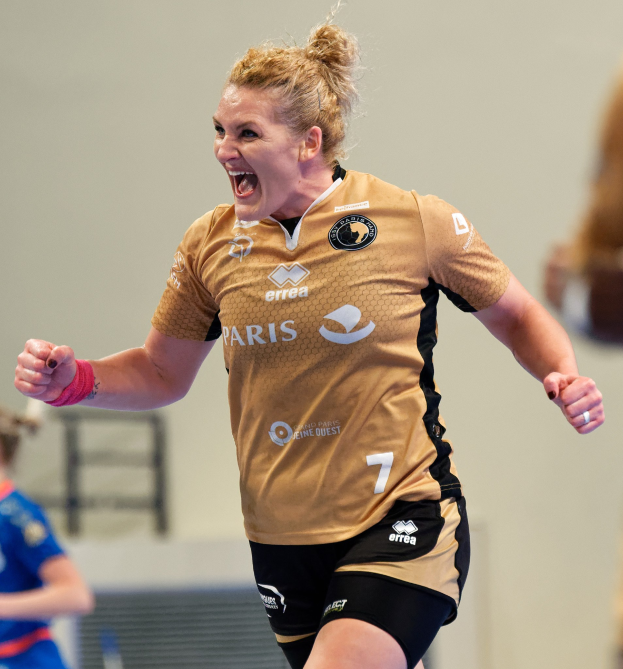 Eine Frau in braunem T-Shirt und schwarzen Shorts rennt und feiert auf einem Futsal-Platz, mit zwei anderen Frauen im Hintergrund.