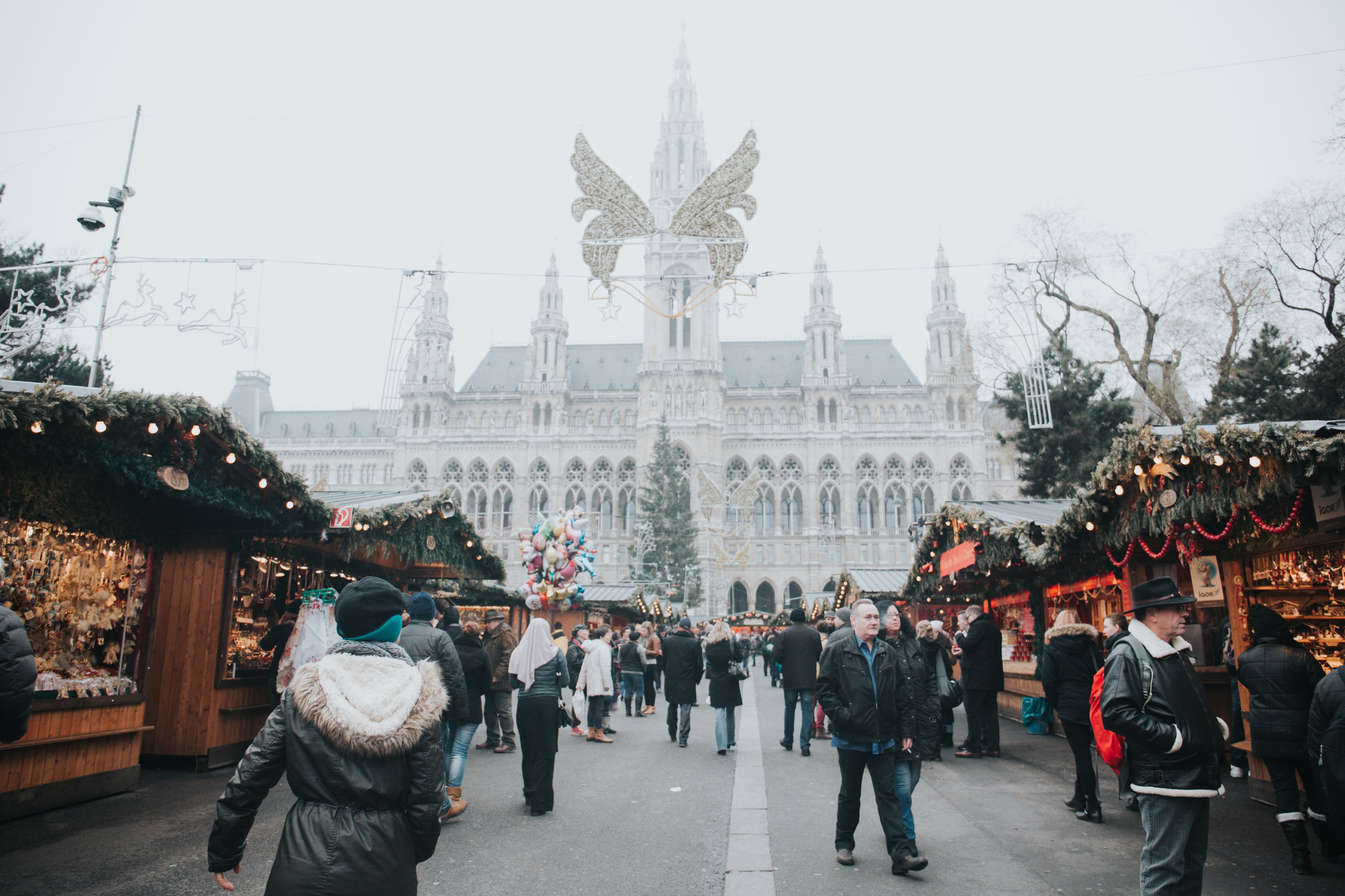 Ein lebendiger Weihnachtsmarkt in Wien, Österreich, mit Menschen, die herumlaufen, Ständen, die mit Lichtern und festlichen Dekorationen geschmückt sind, einem Gebäude mit Fenstern im Hintergrund, Bäumen und einem klaren blauen Himmel.