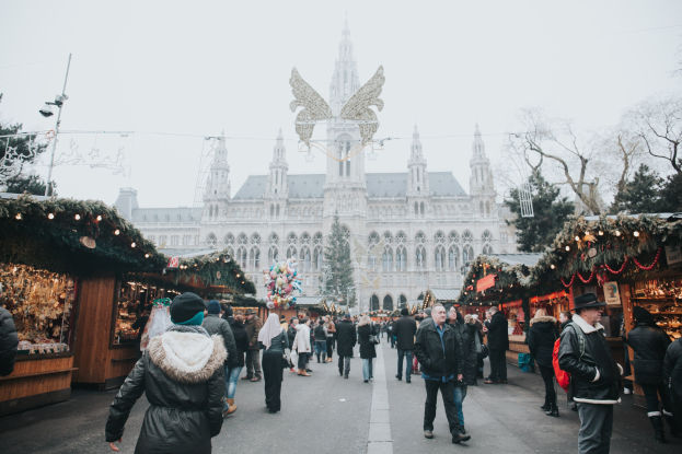 Ein lebendiger Weihnachtsmarkt in Wien, Österreich, mit Menschen, die herumlaufen, Ständen, die mit Lichtern und festlichen Dekorationen geschmückt sind, einem Gebäude mit Fenstern im Hintergrund, Bäumen und einem klaren blauen Himmel.