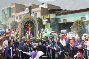 Menschen auf einer Straße vor der Stadt mit einem Kreuz und Blumensträußen in der Mitte, Autos fahren hinter ihnen und Gebäude, Bäume und Straßenlaternen im Hintergrund.