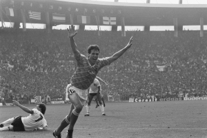 Ein Schwarz-Weiß-Foto eines Fußballspielers, der ein Tor feiert, umgeben von Menschen auf dem Boden, mit Brettern, Fahnen und Pfählen im Hintergrund unter einem klaren Himmel.