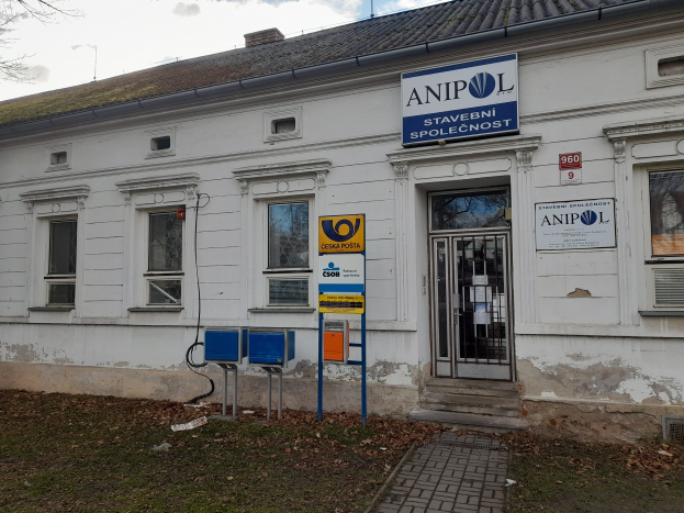 Weißes Gebäude mit "Anipol Post Office"-Schilder, Fenstern, Tür, Weg, Gras, trockenen Blättern, Baum und bewölktem Himmel.
