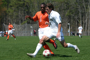 Vier Personen spielen Fußball auf einem Rasenfeld, mit einem Ball im Vordergrund, einige stehen und einige sitzen in der Mitte, sowie Pfosten, Zäune und Bäume im Hintergrund.