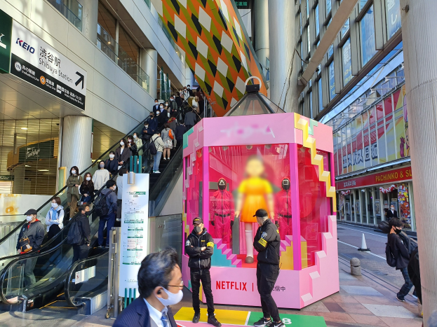 Eine Gruppe von Menschen steht vor einem Gebäude mit Glaswänden und Texttafeln, einige tragen Masken, während hinter einem pinken Kasten mit Puppen und dem Text "Netflix's new Netflix booth in Tokyo" eine Rolltreppe mit Menschen fährt.